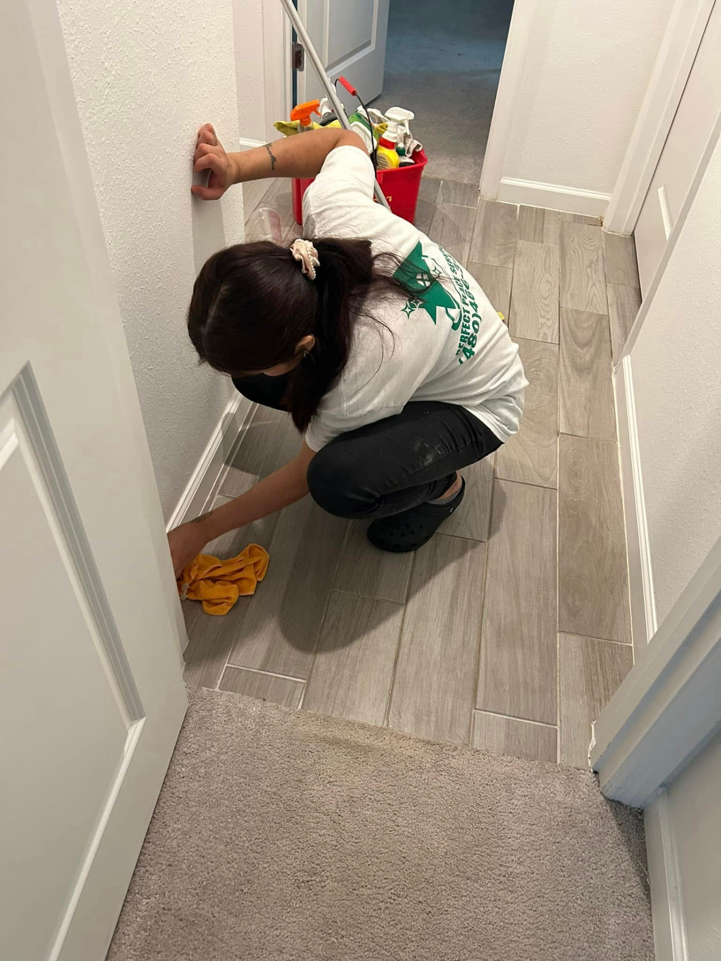 Perfect Place cleaner wiping a hallway baseboard beside a cleaning caddy